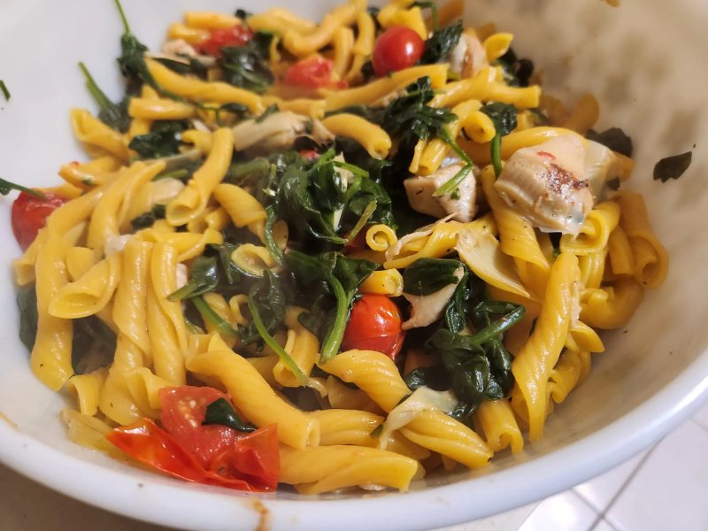 Short twisted pasta with wilted greens, blistered cherry tomatoes and artichoke hearts, in a white bowl.