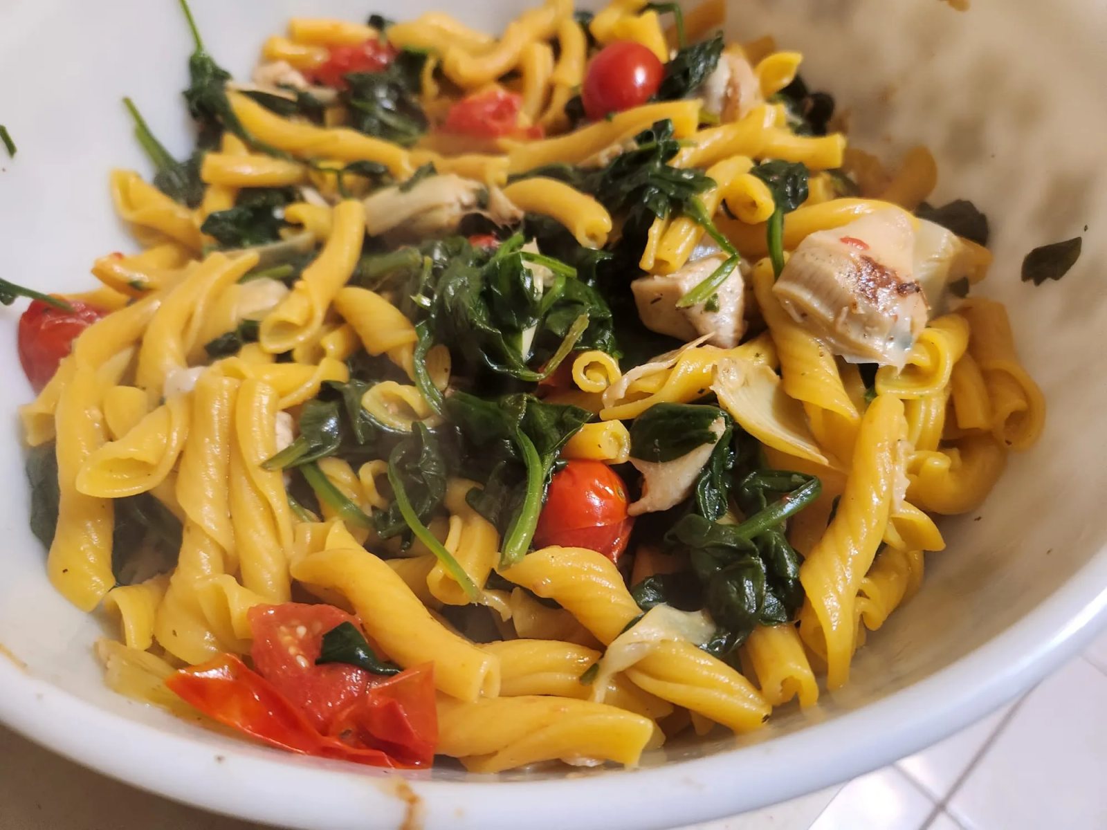 Short twisted pasta with wilted greens, blistered cherry tomatoes and artichoke hearts, in a white bowl.