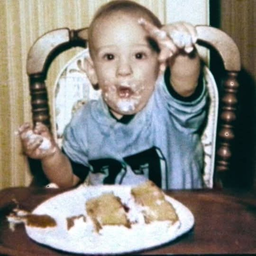 A small boy in a highchair with cake all over his face,
                        one hand raised.